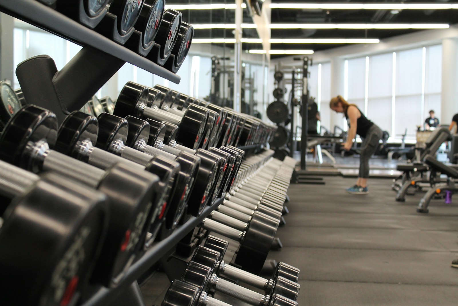 Sala de pesas con rack de mancuernas y persona entrenando al fondo